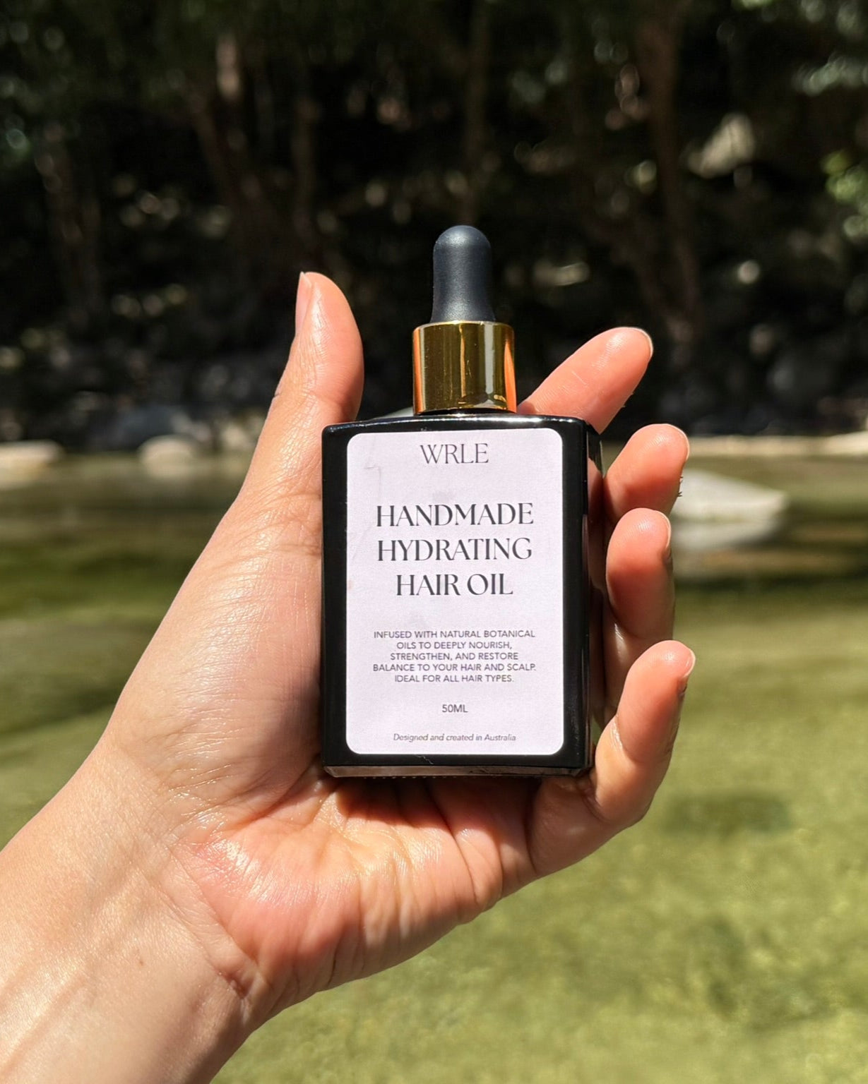 Hand holding a bottle of 'WILF Handmade Hydrating Hair Oil' with a natural background